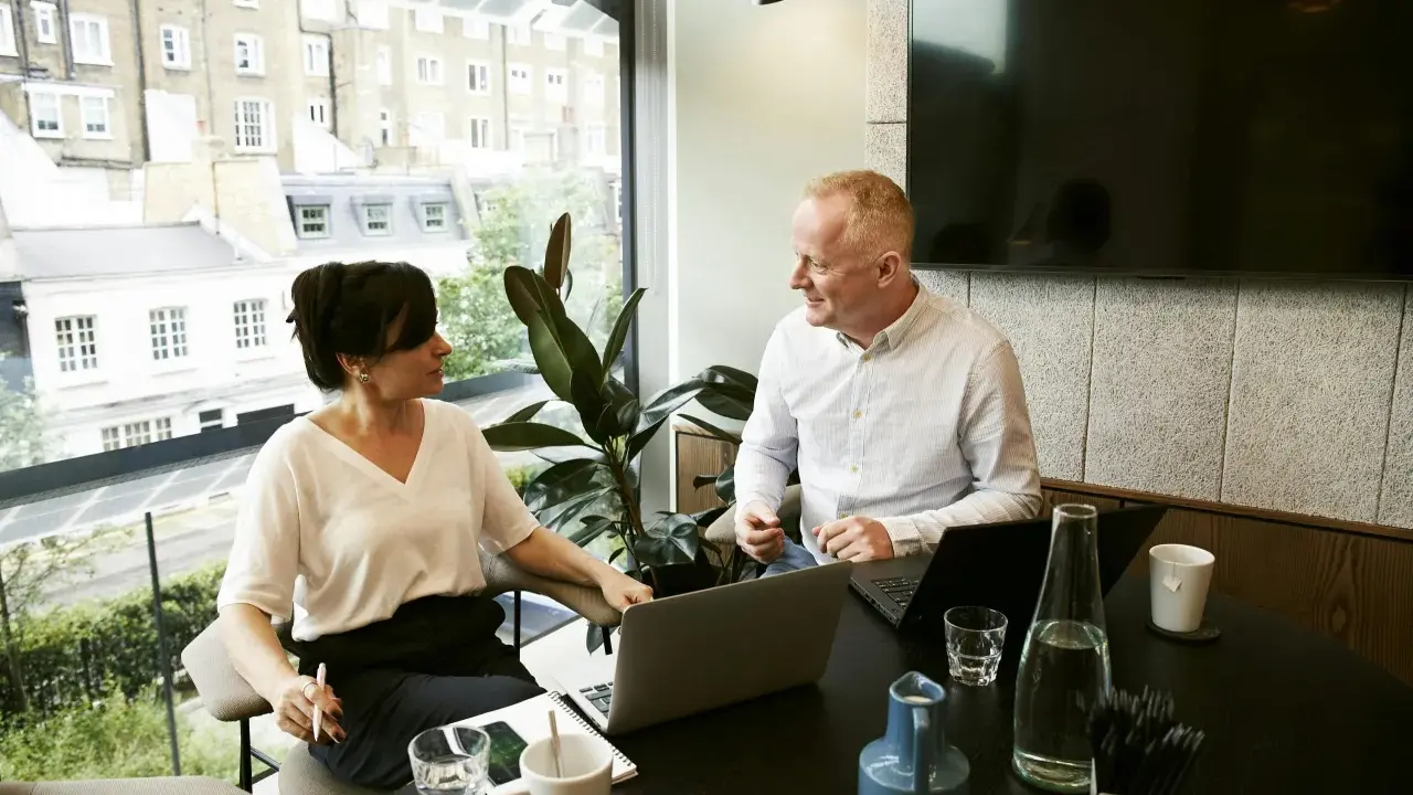 Un hombre y una mujer conversan en una oficina moderna, con portátiles y bebidas. Podría ser una sesión de ceo coaching.