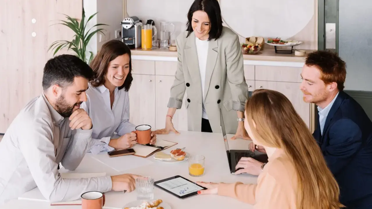 Equipo colaborando en una mesa con café y bocadillos. A pesar de las reuniones largas, el ambiente es positivo y productivo.