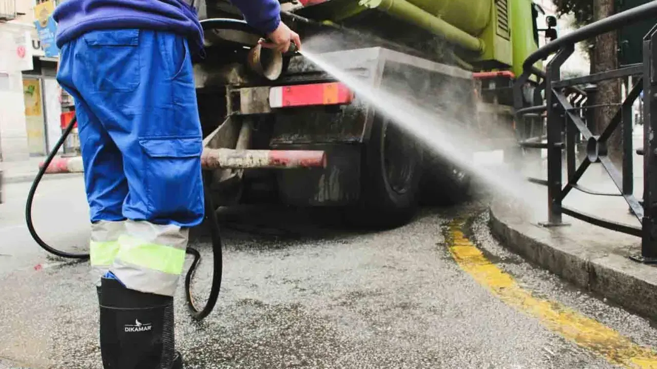Un operario de limpieza, con traje azul y botas negras, rocía agua a presión contra la parte trasera de un camión cisterna verde.