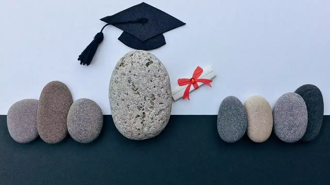 Un birrete de graduación y un diploma con lazo rojo, rodeados de piedras, simbolizan el camino hacia becas MBA y el éxito académico.