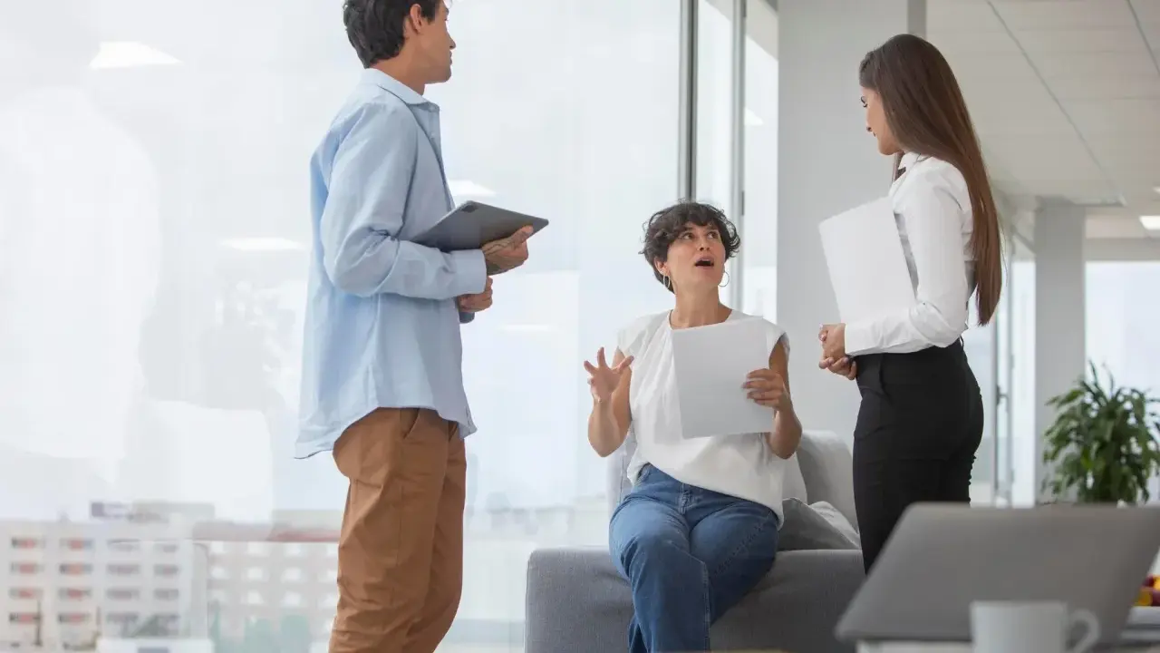 Tres colegas discuten un proyecto. La mujer sentada gesticula mientras explica, buscando cómo mejorar la comunicación interna de una empresa.