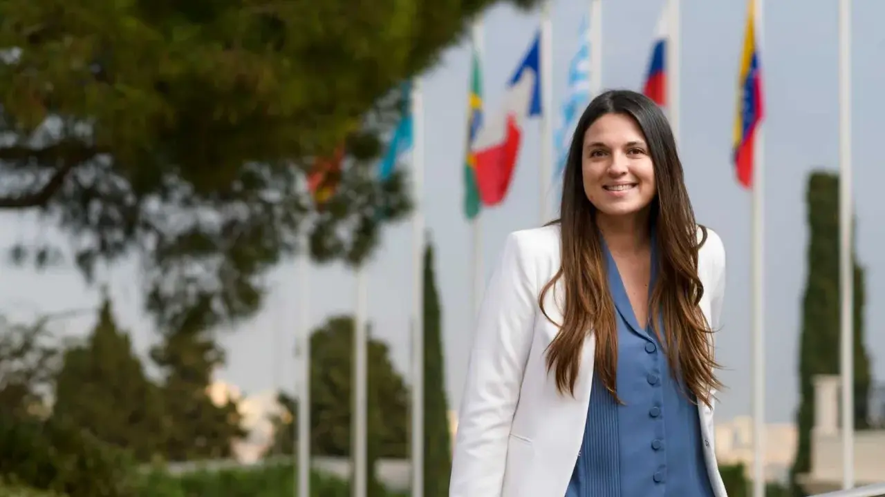 Mujer sonriente con chaqueta blanca y blusa azul, detrás de ella hay banderas. Ella es un pdd.