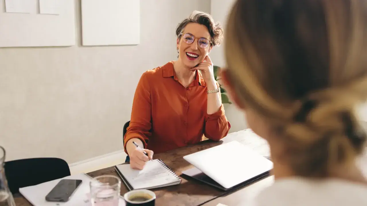 Mujer sonriente con gafas, tomando notas, discutiendo qué podrias aportar a la empresa.