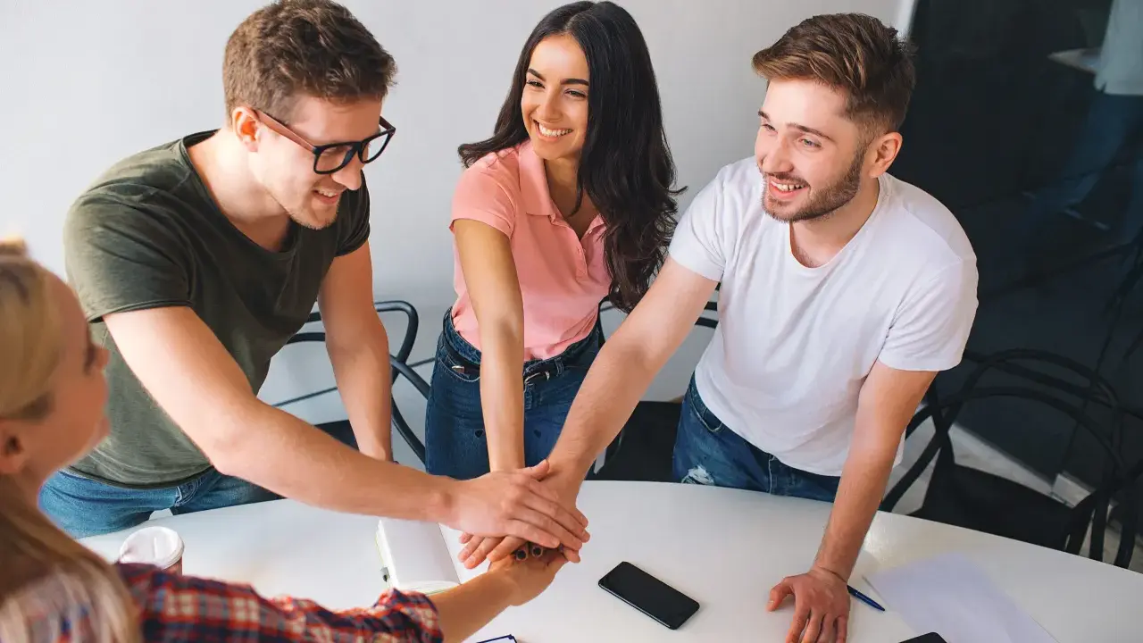 Equipo unido en formación de gestión de equipos. Jóvenes sonríen mientras juntan sus manos en señal de compromiso y colaboración.