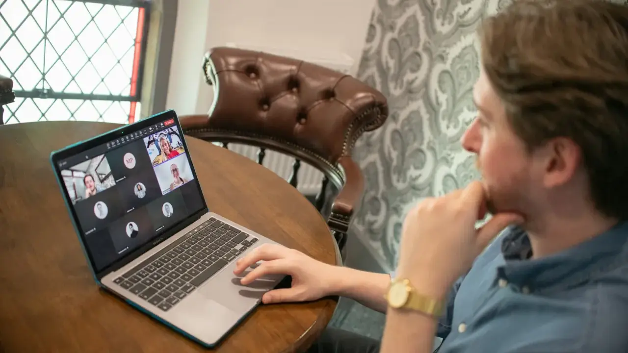 Hombre en videollamada con sus equipos virtuales. Varios rostros aparecen en la pantalla de su portátil, conectados desde diferentes lugares.