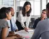 Mujeres y hombres en una reunión discutiendo un proyecto. La líder, con una blusa a cuadros, señala un documento, inspirando un **curso empoderamiento femenino**.