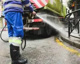 Un operario de limpieza, con traje azul y botas negras, rocía agua a presión contra la parte trasera de un camión cisterna verde.