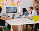 Equipo en una reunión "all hands", discutiendo ideas frente a un monitor y pizarras con notas adhesivas.
