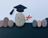 Un birrete de graduación y un diploma con lazo rojo, rodeados de piedras, simbolizan el camino hacia becas MBA y el éxito académico.
