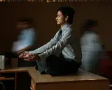 Un joven en posición de loto, con las manos en meditación, busca la calma en medio del ajetreo. Su serenidad contrasta con las figuras borrosas que lo rodean, reflejando la complejidad de la gestión sanitaria.