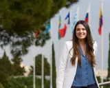 Mujer sonriente con chaqueta blanca y blusa azul, detrás de ella hay banderas. Ella es un pdd.