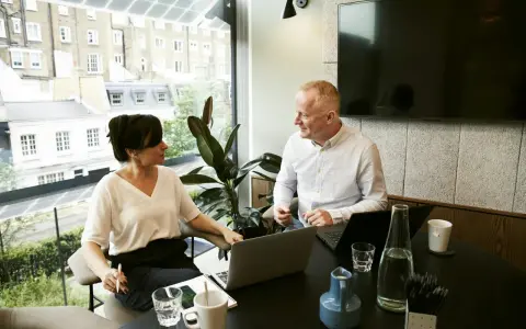 Un hombre y una mujer conversan en una oficina moderna, con portátiles y bebidas. Podría ser una sesión de ceo coaching.