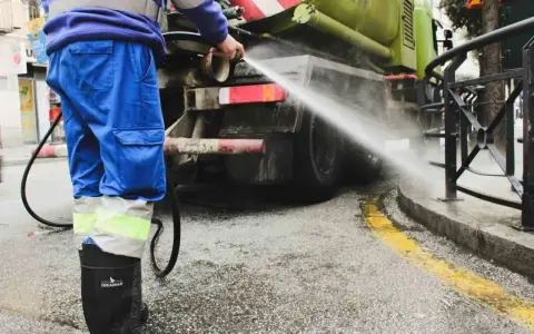 Un operario de limpieza, con traje azul y botas negras, rocía agua a presión contra la parte trasera de un camión cisterna verde.