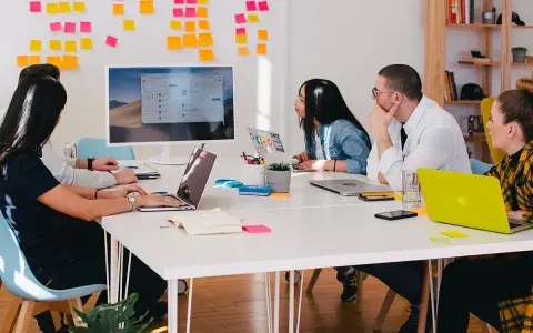 Equipo en una reunión "all hands", discutiendo ideas frente a un monitor y pizarras con notas adhesivas.