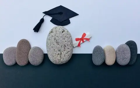 Un birrete de graduación y un diploma con lazo rojo, rodeados de piedras, simbolizan el camino hacia becas MBA y el éxito académico.