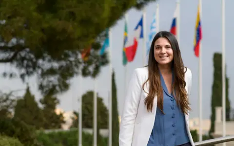 Mujer sonriente con chaqueta blanca y blusa azul, detrás de ella hay banderas. Ella es un pdd.