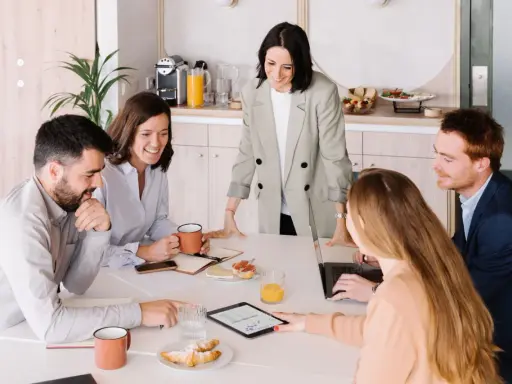 Equipo colaborando en una mesa con café y bocadillos. A pesar de las reuniones largas, el ambiente es positivo y productivo.