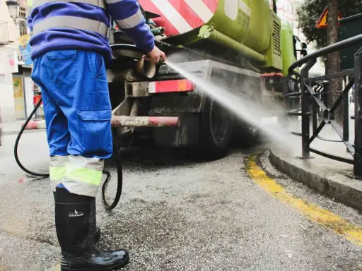 Un operario de limpieza, con traje azul y botas negras, rocía agua a presión contra la parte trasera de un camión cisterna verde.