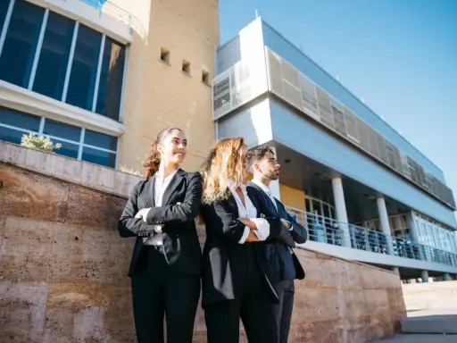 Tres jóvenes profesionales con trajes oscuros, con los brazos cruzados, posan frente a un edificio moderno. Aspiran a estar en los FT MBA rankings.
