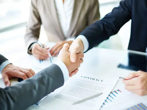 Apertón de manos entre hombres de negocios, con un contrato y gráficos sobre la mesa. Tácticas de negociación exitosas.