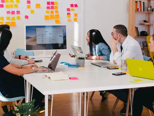 Equipo en una reunión "all hands", discutiendo ideas frente a un monitor y pizarras con notas adhesivas.