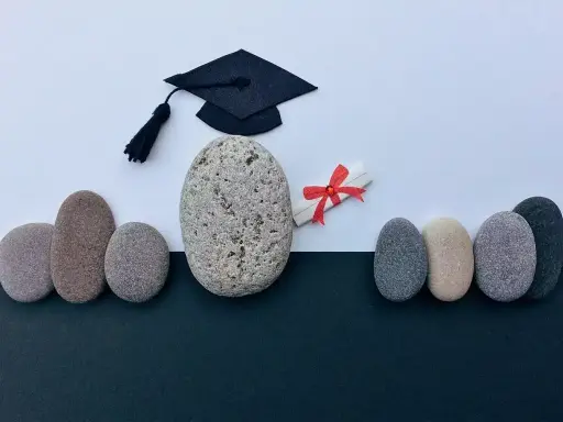 Un birrete de graduación y un diploma con lazo rojo, rodeados de piedras, simbolizan el camino hacia becas MBA y el éxito académico.