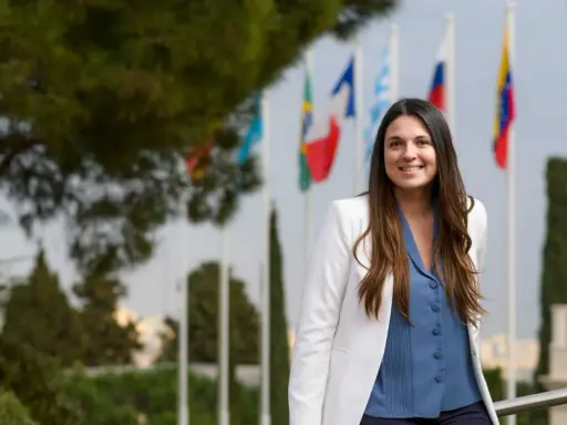 Mujer sonriente con chaqueta blanca y blusa azul, detrás de ella hay banderas. Ella es un pdd.