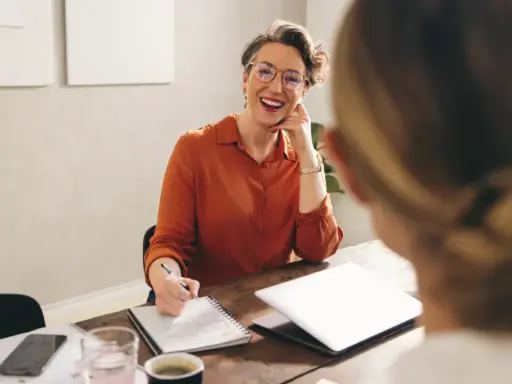 Mujer sonriente con gafas, tomando notas, discutiendo qué podrias aportar a la empresa.