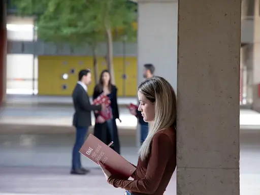 Estudiantes revisan materiales de estudio, como ejemplos de trabajos de oficina, en un campus universitario.