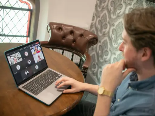 Hombre en videollamada con sus equipos virtuales. Varios rostros aparecen en la pantalla de su portátil, conectados desde diferentes lugares.