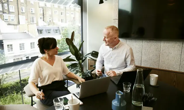 Un hombre y una mujer conversan en una oficina moderna, con portátiles y bebidas. Podría ser una sesión de ceo coaching.