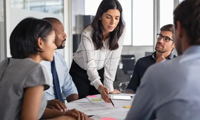 Mujeres y hombres en una reunión discutiendo un proyecto. La líder, con una blusa a cuadros, señala un documento, inspirando un **curso empoderamiento femenino**.