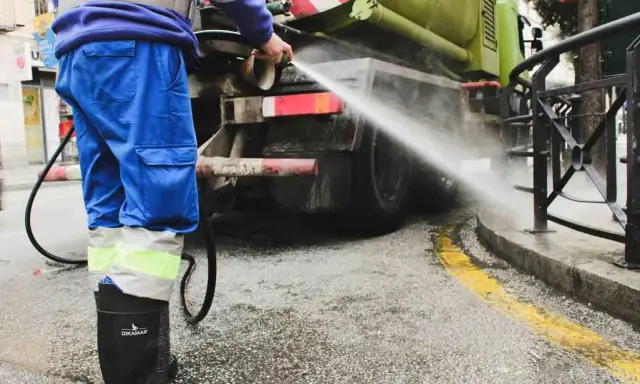 Un operario de limpieza, con traje azul y botas negras, rocía agua a presión contra la parte trasera de un camión cisterna verde.