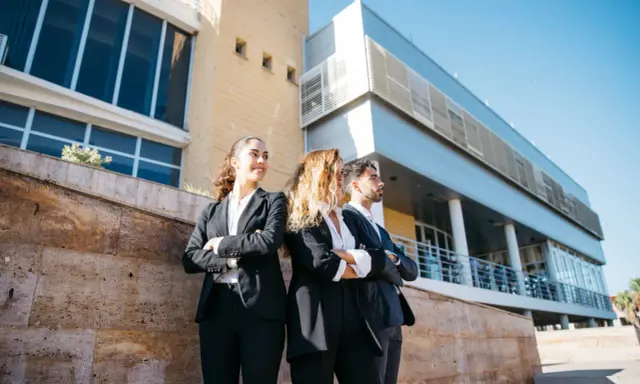 Tres jóvenes profesionales con trajes oscuros, con los brazos cruzados, posan frente a un edificio moderno. Aspiran a estar en los FT MBA rankings.