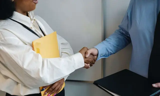 Dos personas se dan la mano, una con una carpeta amarilla, en un momento clave para negociar un salario.
