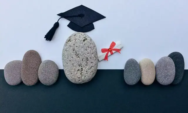 Un birrete de graduación y un diploma con lazo rojo, rodeados de piedras, simbolizan el camino hacia becas MBA y el éxito académico.