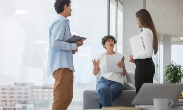 Tres colegas discuten un proyecto. La mujer sentada gesticula mientras explica, buscando cómo mejorar la comunicación interna de una empresa.