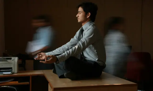 Un joven en posición de loto, con las manos en meditación, busca la calma en medio del ajetreo. Su serenidad contrasta con las figuras borrosas que lo rodean, reflejando la complejidad de la gestión sanitaria.