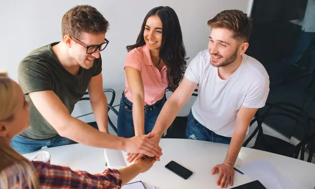 Equipo unido en formación de gestión de equipos. Jóvenes sonríen mientras juntan sus manos en señal de compromiso y colaboración.