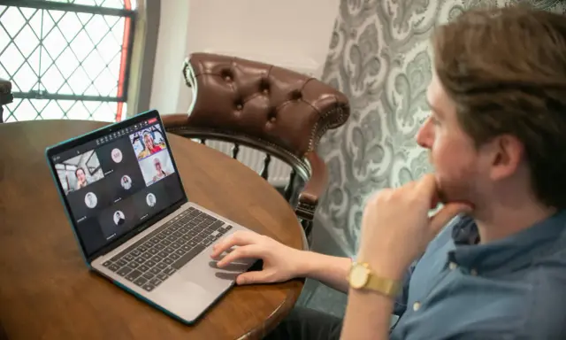 Hombre en videollamada con sus equipos virtuales. Varios rostros aparecen en la pantalla de su portátil, conectados desde diferentes lugares.