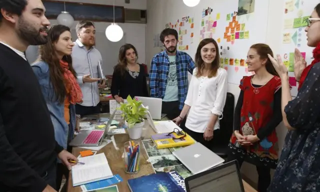 Equipo en una **stand up meeting** discutiendo ideas frente a un tablero lleno de notas adhesivas.