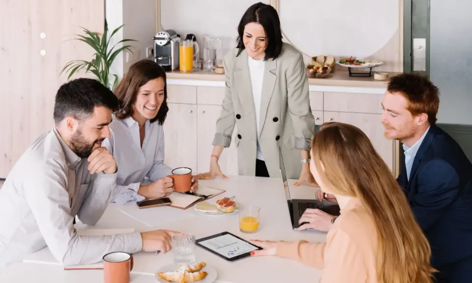 Equipo colaborando en una mesa con café y bocadillos. A pesar de las reuniones largas, el ambiente es positivo y productivo.