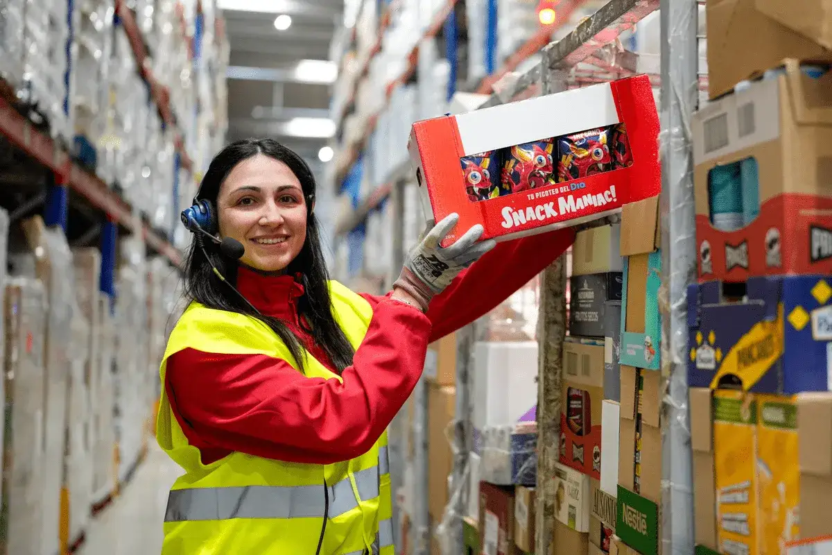 Mujer con chaleco reflectante y auriculares, sonr&iacute;e mientras coloca una caja de snacks en un almac&eacute;n. &iexcl;Un d&iacute;a m&aacute;s en su curso de reponedor!