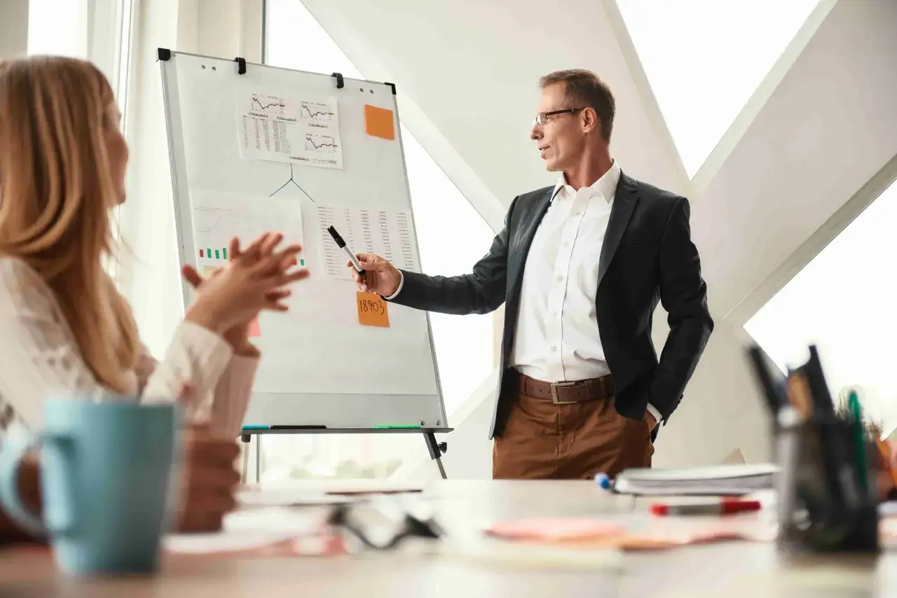 Un hombre en traje da una presentaci&oacute;n de coaching ejecutivo, se&ntilde;alando un gr&aacute;fico en un rotafolio a una mujer.