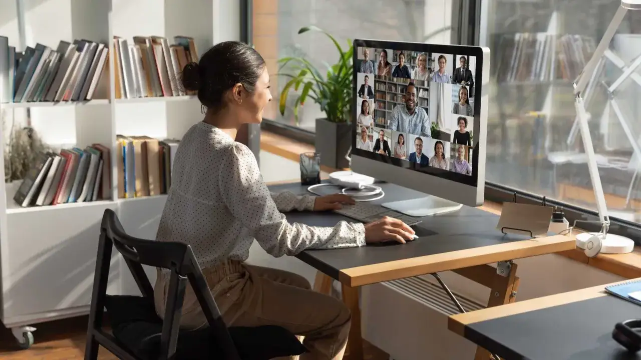 Mujer en videollamada, gestionando equipos a distancia. En pantalla, rostros de colegas en una cuadr&iacute;cula.