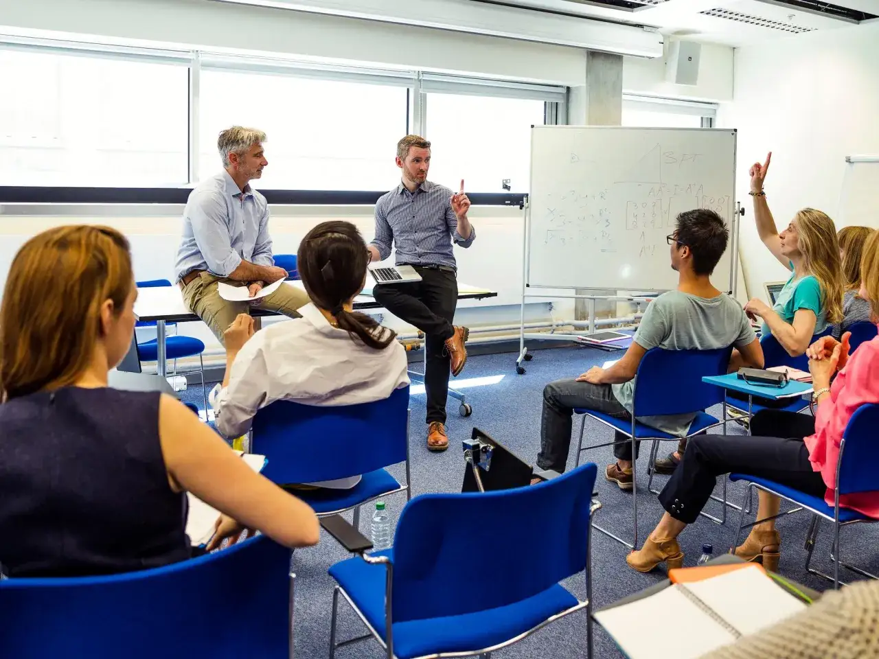 Un profesor explica un concepto en una clase de MBA Master. Los estudiantes participan activamente, uno levanta la mano.