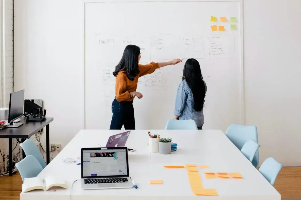 Dos mujeres discuten un proyecto en una pizarra. Una excelente oferta de pr&aacute;cticas para aprender y colaborar.