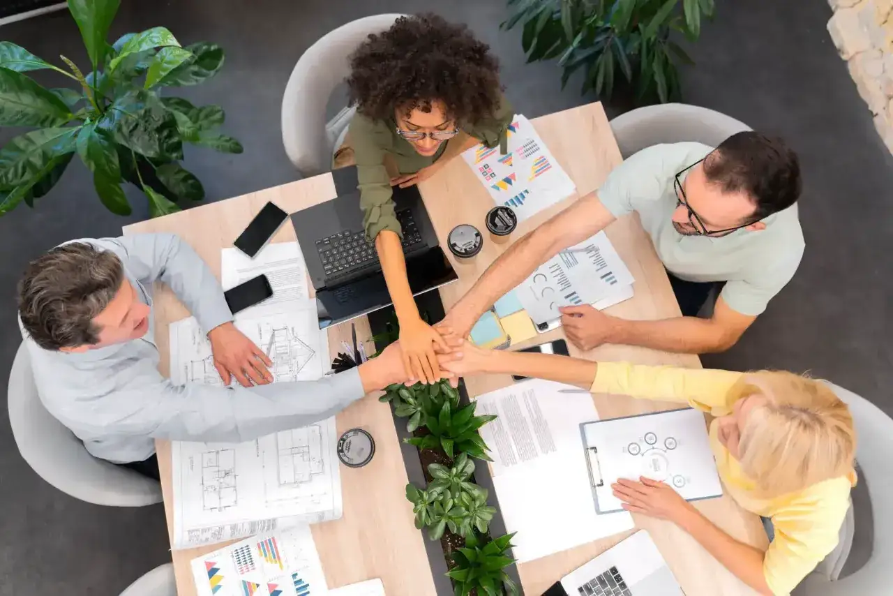 Equipo unido en reuniones de empresa, manos entrelazadas sobre la mesa con gr&aacute;ficos y planos.
