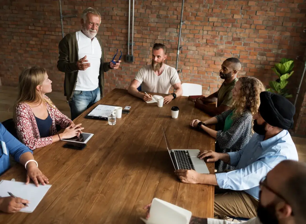 Un grupo diverso de personas participa en reuniones largas alrededor de una mesa de madera, discutiendo ideas y colaborando en proyectos.