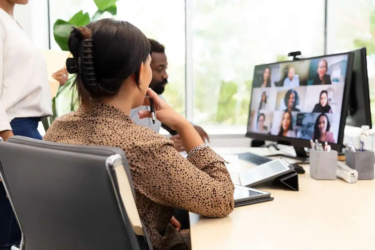 Personas en una oficina participando en reuniones virtuales. La pantalla muestra una cuadrícula de rostros sonrientes, conectando equipos a distancia.